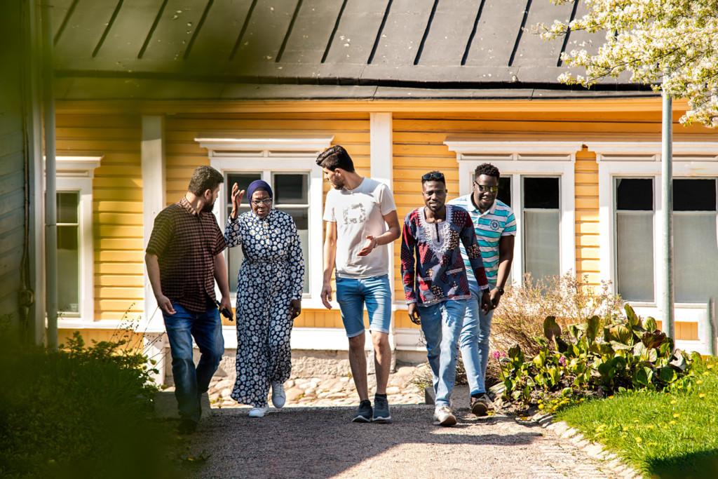 En grupp invandrare på gården med ett trähus på bakgrund.