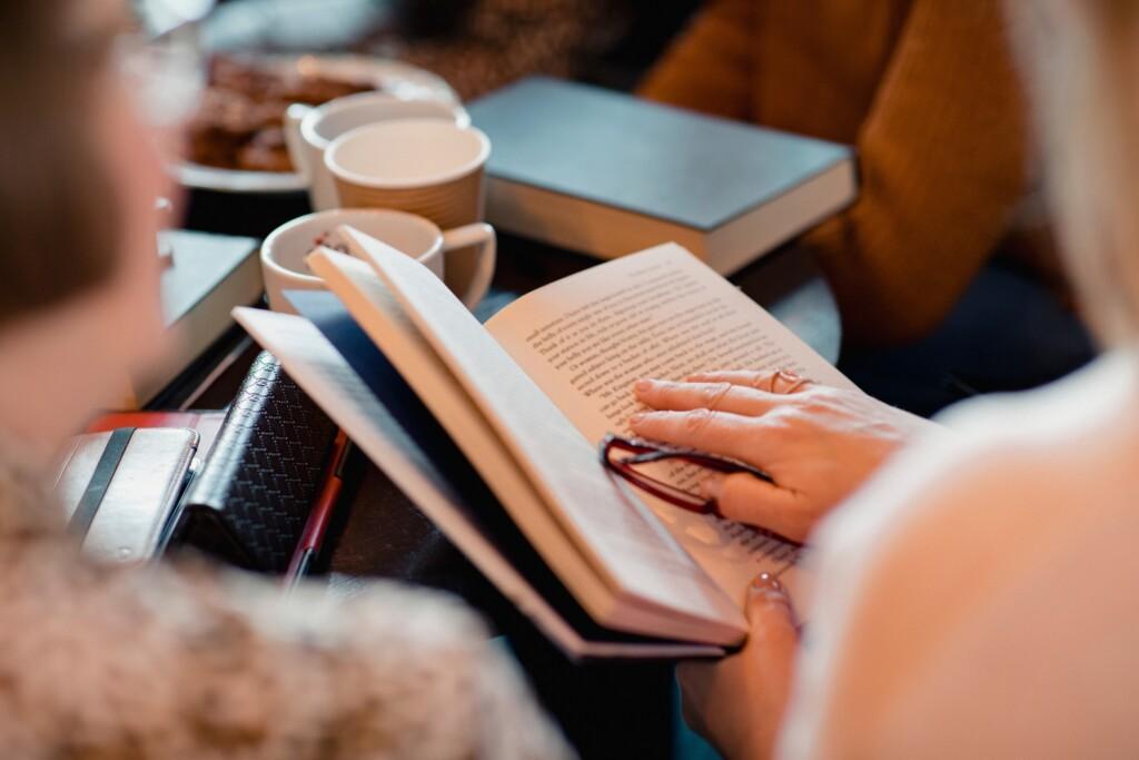 Kaffekoppar på bordet och en person håller en bok, andra människor på bakgrunden också.