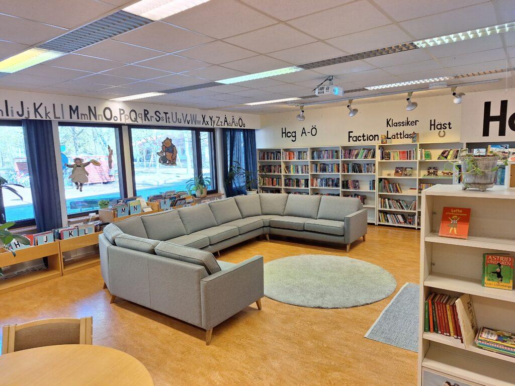 En soffa och hyllor iett biblioteksutrymme med fönster. På väcken löper alfabeterna och titlarna för hyllorna.