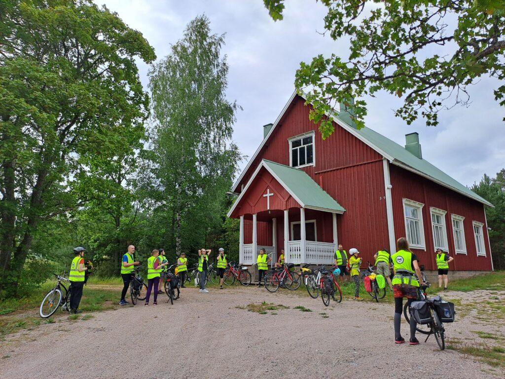 Keltaliivisiä pyöräilijöitä kokoontuneena puisen kirkkorakennuksen ympärille.