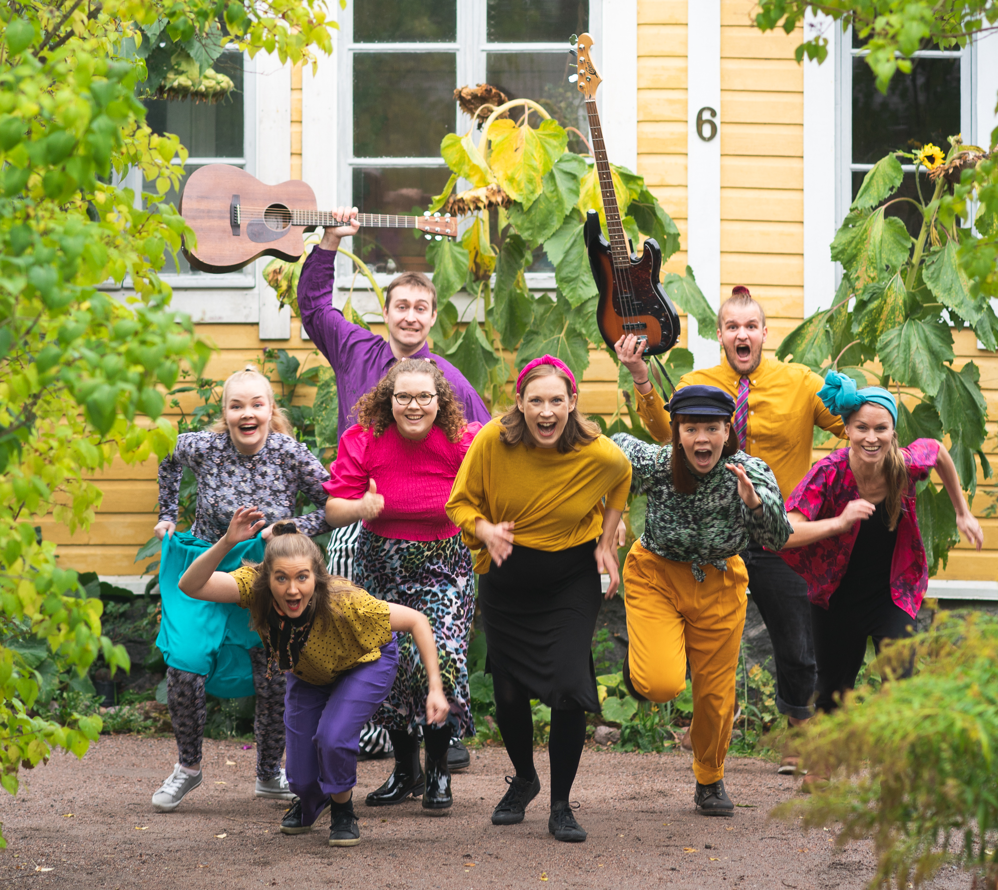 Kahdeksan värikkäästi pukeutunutta muusikkoa juoksee puutarhassa soittimet kädessään