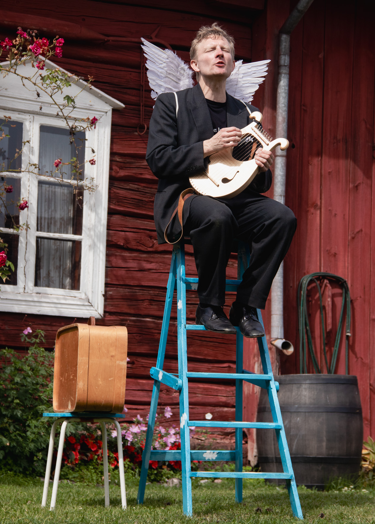 Mies istuu tikkailla punaisen puutalon edessä. Miehellä on siivet selässä.